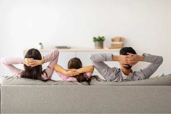 Neruofeedback: Drei Personen sitzen nebeneinander auf einem grauen Sofa, mit dem Rücken zur Kamera. Links eine Frau mit langen dunklen Haaren, in der Mitte ein Kind mit dunklen, zusammengebundenen Haaren, rechts ein Mann mit kurzen dunklen Haaren. Alle haben die Hände hinter dem Kopf verschränkt. Im Hintergrund eine helle Wand mit Sideboard und Pflanze.