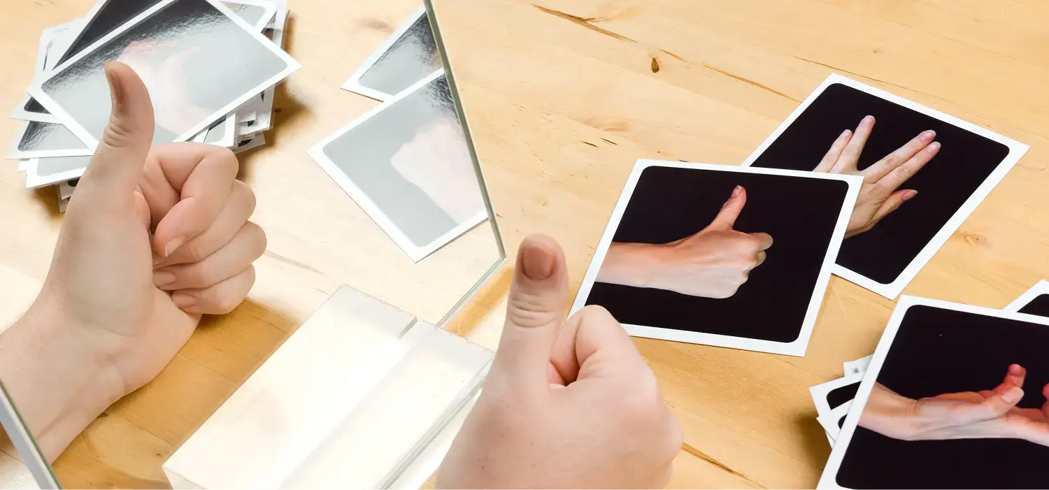 Neurologie Ergotherapie: Zwei Hände liegen auf einem Tisch, dazwischen ein rechteckiger Spiegel. Im Spiegelbild ist eine der Hände doppelt zu sehen. Auf dem Tisch liegen karten mit Handstellungen.