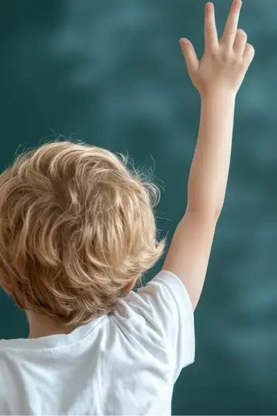 Pädiatrie Ergotherapie: Kind mit kurzen, hellbraunen Haaren sitzt auf einem Stuhl, Rücken zur Kamera. Der rechte Arm ist erhoben, die Hand zeigt nach oben. Hintergrund unscharf und hell.