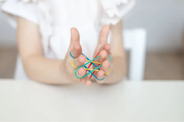 Ergotherapie-SonjaWandel-Stuttgart-Paedieatrie-Haende Pädiatrie Ergotherapie: Kind sitzt an einem weißen Tisch, nur Oberkörper und Arme sind sichtbar. Beide Hände halten mehrere bunte Gummibänder (gelb, blau, grün, rot), die zwischen den Fingern gespannt sind und ein Netz bilden.