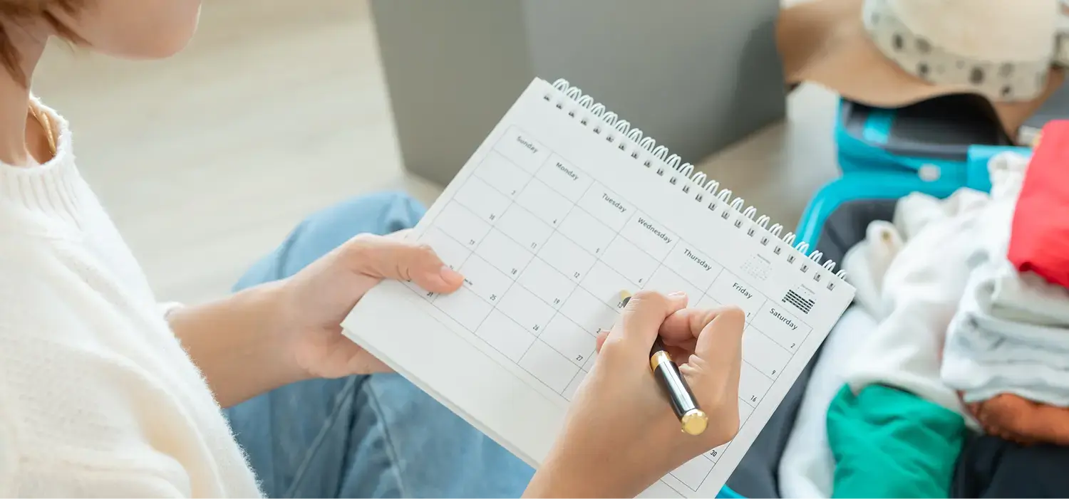 Psychiatrie Ergotherapie: Eine Person hält einen aufgeschlagenen Tischkalender mit Monatsübersicht. Mit der rechten Hand wird ein Datum markiert. Links im Bild sind die Beine in blauer Jeans sichtbar, rechts daneben ein Koffer mit Kleidung.