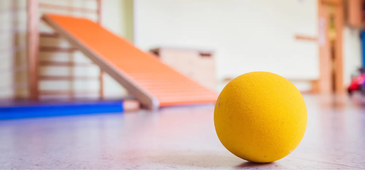 Ergotherapie: Gelber Ball liegt auf einem blau-orangenen Boden. Im Hintergrund eine unscharfe Rampe und weiße Wand.