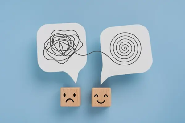 Psychologie Ergotherapie: Zwei quadratische Würfel mit Gesichtern hängen nebeneinander an der Wand. Links ein trauriges Gesicht mit schwarzer, wirrer Linie darüber; rechts ein lächelndes Gesicht mit ordentlicher, spiralförmiger Linie darüber. Hintergrund hellblau.