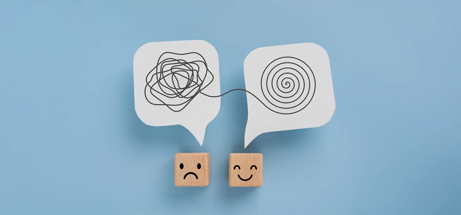 Psychologie Ergotherapie: Zwei quadratische Würfel mit Gesichtern hängen nebeneinander an der Wand. Links ein trauriges Gesicht mit schwarzer, wirrer Linie darüber; rechts ein lächelndes Gesicht mit ordentlicher, spiralförmiger Linie darüber. Hintergrund hellblau.