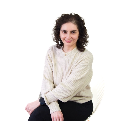 Derya Müller: Frau mit lockigen dunklen Haaren, trägt hellen Pullover, sitzt seitlich auf einem weißen Stuhl.