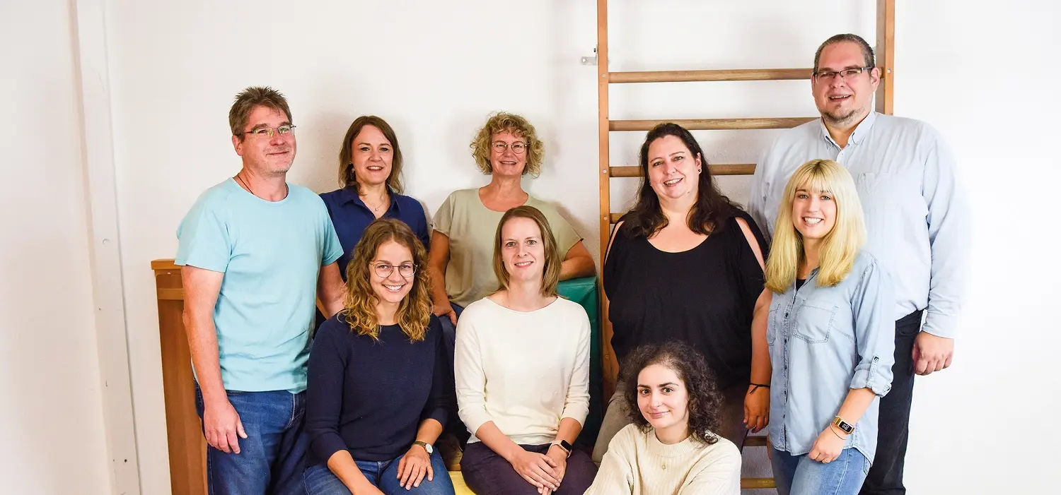 Ergotherapie Praxis Sonja Wandel Team: Neun Personen stehen und sitzen in zwei Reihen vor einer hellen Wand. Hinten vier Frauen und zwei Männer, vorne drei Frauen. Links im Hintergrund ist eine Holz-Sprossenwand sichtbar. Alle schauen in die Kamera und lächeln.