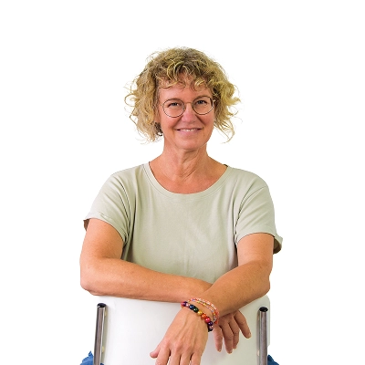 Kerstin Steckroth: Frau mit lockigen blonden Haaren, trägt hellgrünes Shirt und Brille, lehnt an einer Stuhllehne.