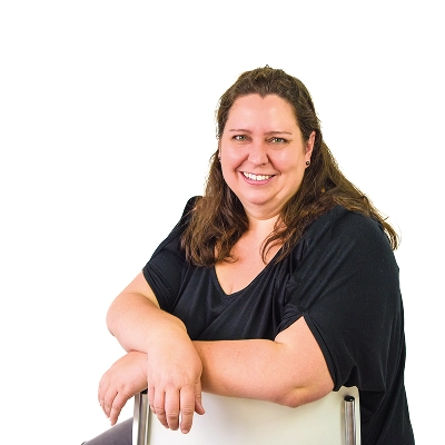 Sonja Wandel: Frau mit langen dunklen Haaren, trägt schwarzes Oberteil, lehnt lächelnd an einer weißen Stuhllehne.