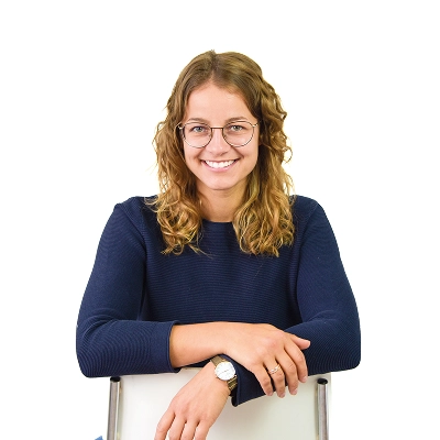 Sophia Thieß: Frau mit langen blonden Haaren und Brille, trägt dunkles Oberteil, lehnt auf einer weißen Stuhllehne.